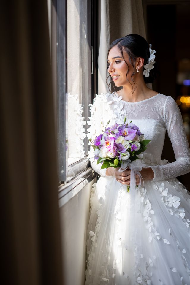 Mariée près d'une fenêtre, un bouquet de fleurs mauves dans les mains
