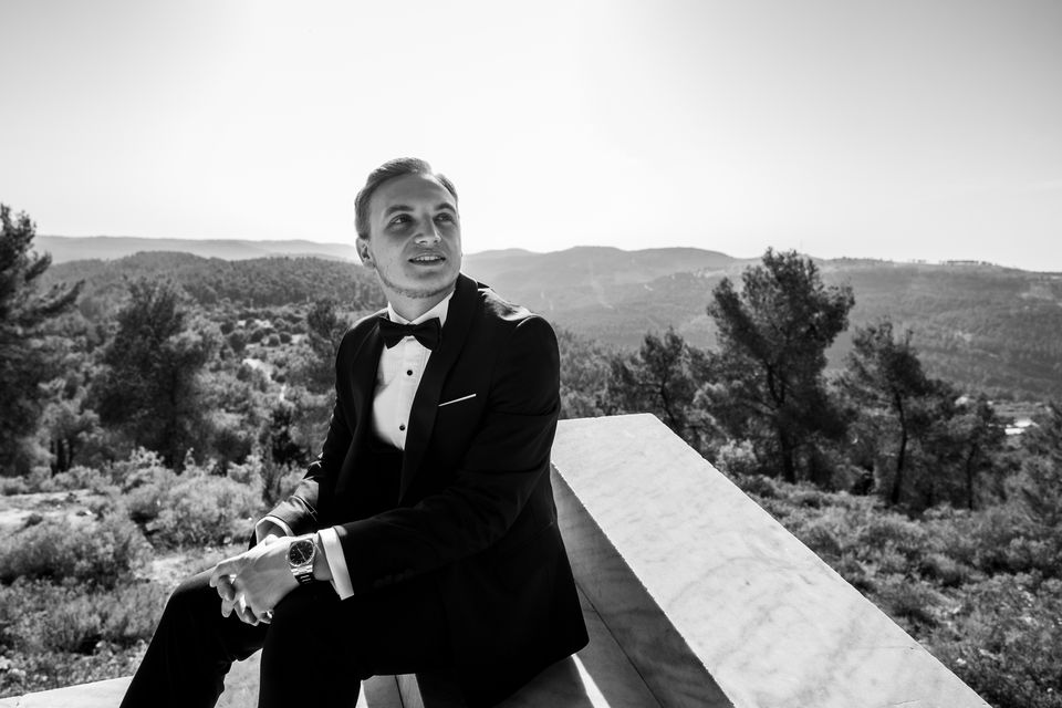 Marié en smoking assis sur un muret, face aux collines de Judée, en noir et blanc