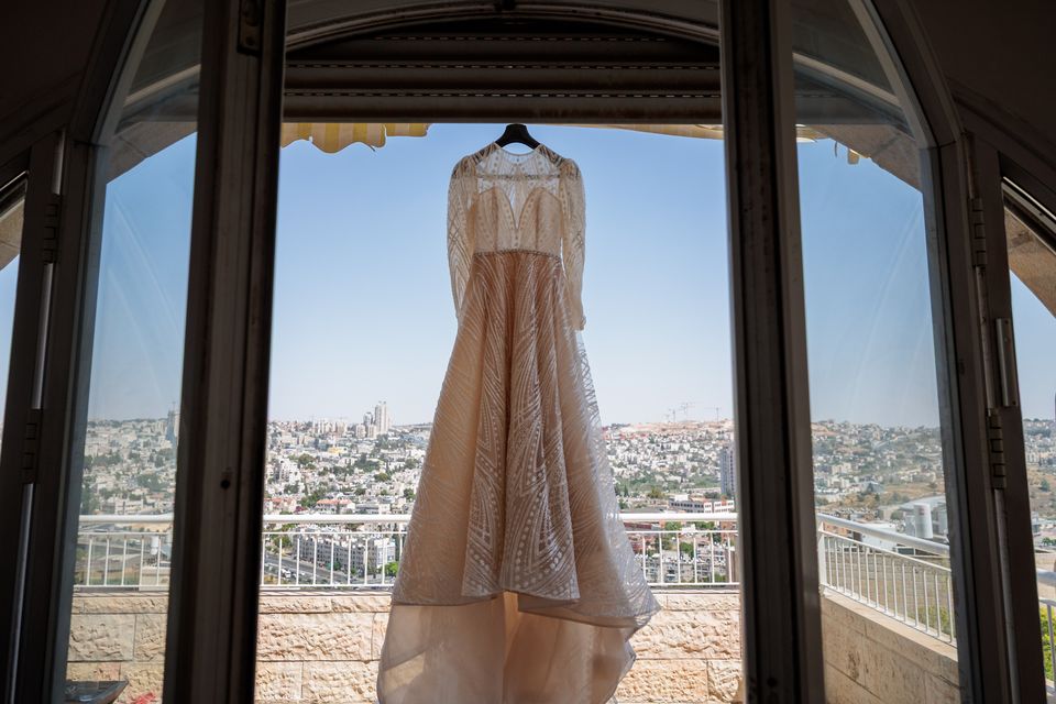Robe de mariée suspendue dans une fenêtre en arc, avec la ville de Jeusalem en arrière-plan